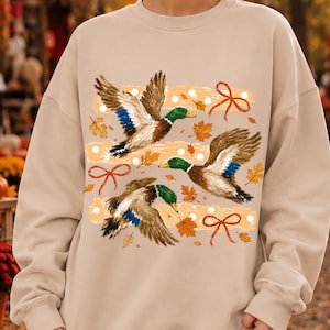 May include: A sand-colored crewneck sweatshirt featuring a graphic print of three mallard ducks in flight. The design includes scattered orange autumn leaves, white polka dots, and orange ribbon bows arranged in horizontal bands across the chest.