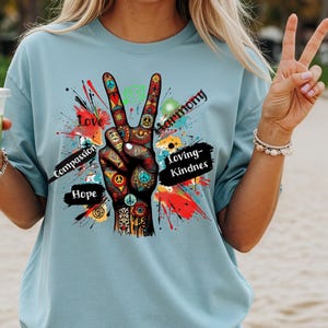 May include: A light blue t-shirt featuring a colorful peace sign graphic. The graphic includes the words "Love", "Compassion", "Hope", "Harmony", and "Loving-Kindness". The peace sign is decorated with various symbols and patterns.