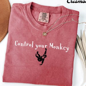 May include: A red t-shirt with the phrase "Control your Monkey" in white text and a black monkey graphic. The shirt has a crew neck and short sleeves. A gold necklace with a pendant is draped across the shirt.