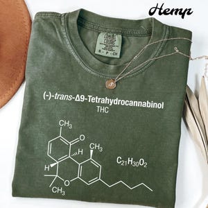 May include: An olive green t-shirt featuring white text and a molecular structure diagram. The text includes "(-)-trans-Δ9-Tetrahydrocannabinol THC" and "C21H30O2". A gold necklace with a pendant is draped across the shirt. The word "Hemp" is written in cursive.