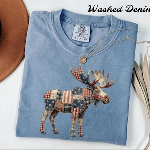 May include: Light blue t-shirt with a moose design. The moose is made of patchwork fabric with red, white, and blue patterns. The shirt has the words "Washed Denim" printed at the top. A gold necklace is draped across the shirt.