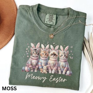 May include: Moss green Comfort Colors t-shirt featuring four cats wearing bunny ears and sweaters, with a small stuffed bunny. The text "Meowy Easter" is printed below the cats. A silver necklace with a pendant is draped across the shirt.