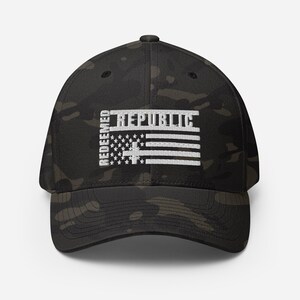 Puede incluir: Gorra de béisbol de camuflaje negro y verde. La gorra presenta el texto "REDEEMED REPUBLIC" en blanco, con un diseño de bandera blanca. El sombrero tiene una visera curva y un ajuste estructurado.