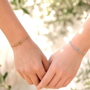 Könnte beinhalten: Zwei dünne Armbänder, eines in poliertem Goldton und eines in poliertem Silberton, jeweils mit einem zarten Knotenmotiv in der Mitte. Die Armbänder bestehen aus einem doppelsträngigen Band, das am Knoten zusammenläuft und so wirkt.