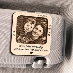 Könnte beinhalten: Ein quadratischer, heller Holz-Clip für die Sonnenblende mit einem eingravierten Foto eines Kindes und eines Erwachsenen. Oben steht der Text 10.11.2025 und unten der Text Bitte fahre vorsichtig, wir brauchen Dich hier bei uns.