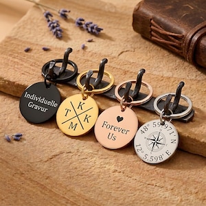 May include: A selection of engraved keyrings in different metallic finishes. One black keyring has the text "Individuelle Gravur". A gold keyring has initials. A rose gold keyring has a heart and the words "Forever Us". A silver keyring has coordinates.