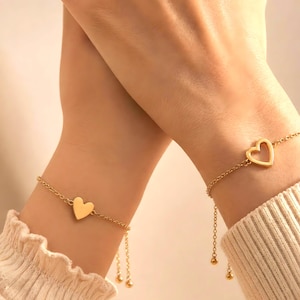 Könnte beinhalten: Zwei goldfarbene Armbänder mit Herzmotiven. Eines hat einen soliden Herzanhänger, das andere eine herzförmige Aussparung. Beide haben zarte Kettenglieder und baumelnde Kugelakzente, die ein schlichtes, aber elegantes Design schaffen.