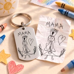 Könnte beinhalten: Ein rechteckiger Schlüsselanhänger aus silberfarbenem Metall mit einer Gravur einer Kinderzeichnung, die eine Person mit Krone und das Wort MAMA darüber zeigt. Das Design entspricht einem zerknitterten weißen Papier mit der gleichen Zeichnung.