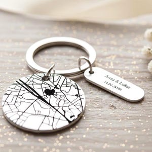 May include: A silver keyring with a circular pendant displaying a black and white map design and a heart. A smaller rectangular tag is attached, with the engraved text "Anna & Lukas 14.02.2022".