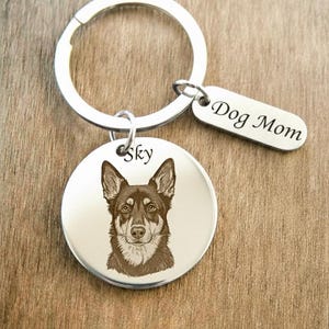 Könnte beinhalten: Ein silberfarbener Schlüsselanhänger mit einem runden Anhänger, der eine braune Hundeillustration und den Namen Sky zeigt, sowie einem kleinen rechteckigen Anhänger mit der Gravur Dog Mom, befestigt an einem runden Schlüsselring.