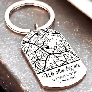 Peut inclure: Un porte-clés en métal doré rose avec une plaque rectangulaire aux coins arrondis, gravée d'un plan de ville noir, d'un petit cœur, et du texte "Wo alles begann", "52.372585°, 9.732737°" ainsi que les prénoms "Gaby & Axel".