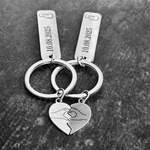 Puede incluir: Dos llaveros plateados con un colgante de corazón, cada mitad con manos formando un corazón. Los llaveros tienen etiquetas rectangulares con la fecha "10.08.2025" y un diseño de nube. Los llaveros están conectados entre sí.