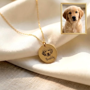 Könnte beinhalten: Goldfarbene Halskette mit rundem Anhänger, graviert mit dem Gesicht eines Golden Retrievers und dem Namen "Buddy". Im Hintergrund befindet sich ein gerahmtes Foto eines Golden Retriever-Welpen. Das Schmuckstück liegt auf weißem Stoff.