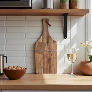 Könnte beinhalten: Holzschneidebrett mit Griff, verziert mit einem Kranz und Initialen. Ein Etikett mit Text ist mit Bindfaden an der Platte befestigt. Ein Glas Weißwein und eine Schüssel mit Nüssen stehen auf der Theke.
