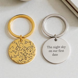 Könnte beinhalten: Zwei Schlüsselanhänger, einer goldfarben und einer silberfarben, jeweils mit einem runden Anhänger. Der goldfarbene Schlüsselanhänger zeigt ein Sternbilddesign. Der silberne Schlüsselanhänger hat den Text "The night sky on our first date".