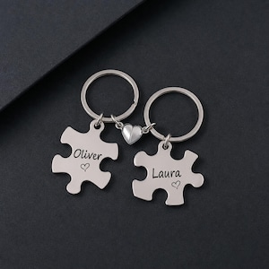 Op de afbeelding: Twee zilverkleurige metalen puzzelstuk-sleutelhangers verbonden door een kleine hartvormige bedel. Het ene stuk is gegraveerd met de naam Oliver en een klein hartje, het andere met de naam Laura en een klein hartje. Elk stuk heeft een ring.