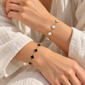 Könnte beinhalten: Zwei goldfarbene Kettenarmbänder mit einer Reihe von vierblättrigen Kleeblattmotiven. Ein Armband zeigt weiße Kleeblatt-Anhänger, während das andere Armband schwarze Kleeblatt-Anhänger aufweist. Beide Schmuckstücke sind filigrane Gliederarmbänder.
