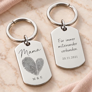 Op de afbeelding: Twee zilverkleurige metalen sleutelhangers in de vorm van een dog tag met ringen. Eén hanger bevat het woord Mama, een hartvormige vingerafdruk en de letters M & E. De andere hanger toont de Duitse tekst Für immer miteinander verbunden en 20.11.2021.
