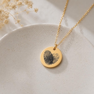 Könnte beinhalten: Eine goldfarbene Halskette mit einem runden Anhänger, der einen schwarzen Fingerabdruck in Herzform zeigt. Der Anhänger ist an einer zarten goldfarbenen Kette befestigt und bildet ein schlichtes, elegantes Schmuckstück mit persönlicher Note.