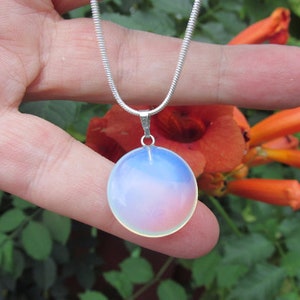 May include: A round, iridescent pendant necklace with a silver chain. The pendant has a soft, opalescent glow, transitioning from white to pink and blue. The background features orange flowers and green foliage.