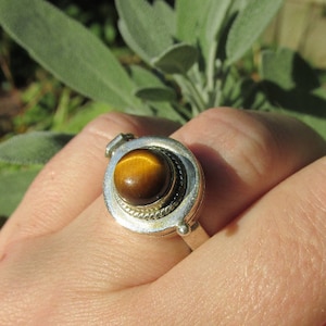 Könnte beinhalten: Ein silberner Ring mit einem runden, braunen Tigerauge-Edelstein, der in einer runden Fassung sitzt. Der Ring hat ein Scharnierdesign, das es ermöglicht, die Fassung zu öffnen und zu schließen.