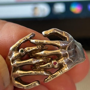 Op de afbeelding: Een zilveren skelethandring met drie rode en groene edelstenen. De ring is ontworpen om eruit te zien als twee handen die elkaar vasthouden.