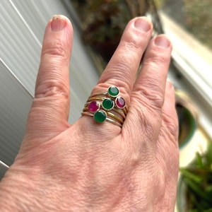 May include: A set of five gold rings with green and red gemstones. The rings are stacked on a finger.