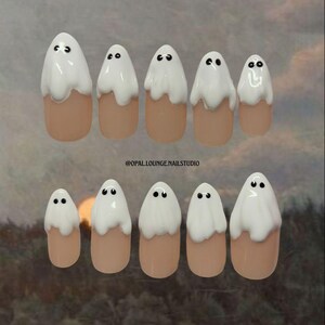 以下が含まれることがあります： ハロウィーンをテーマにしたつけ爪のセット。各爪はヌードベースで塗装され、白いゴーストデザインが施されています。ゴーストは黒い点の目を持っています。爪の先端は楕円形です。