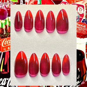May include: Set of oval-shaped, press-on nails with a gradient design. The nails transition from a deep red at the base to a lighter pink at the tips. The background features Coca-Cola branding.