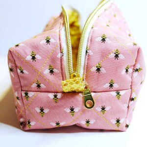 May include: Pink quilted cosmetic bag with a repeating pattern of yellow and black bees. The bag has a gold zipper and a yellow and white interior. The zipper pull is a patterned fabric.