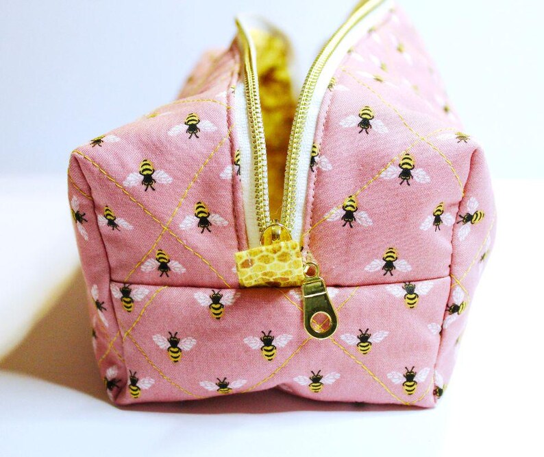 May include: Pink quilted cosmetic bag with a repeating pattern of yellow and black bees. The bag has a gold zipper and a yellow and white interior. The zipper pull is a patterned fabric.