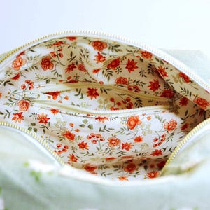 May include: Open view of a light green bag with a white zipper. The interior features a floral pattern with red flowers and green leaves on a white background. The bag's exterior has a daisy pattern.