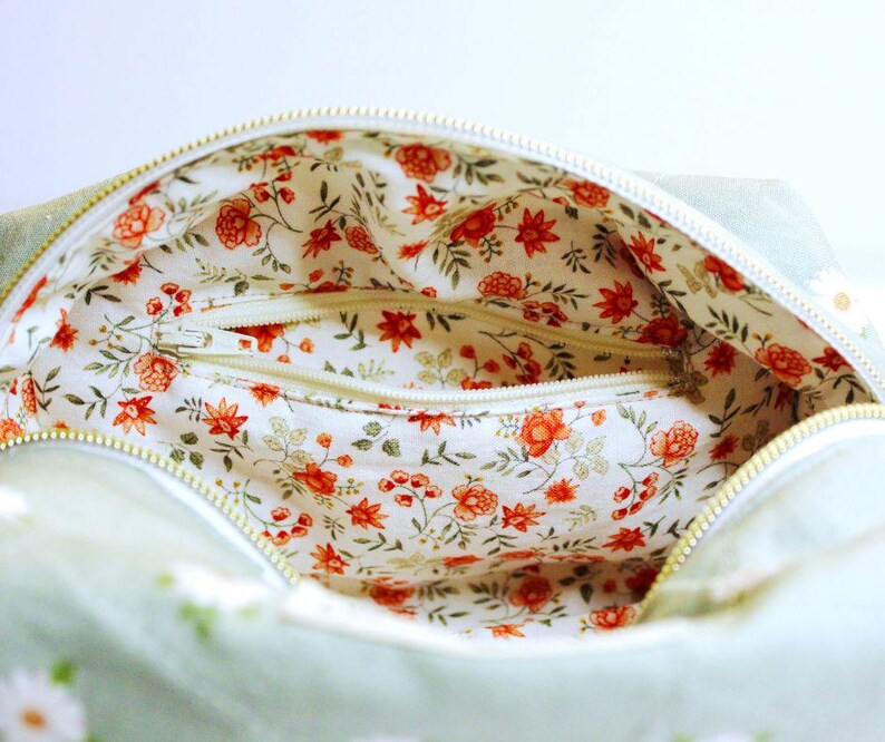 May include: Open view of a light green bag with a white zipper. The interior features a floral pattern with red flowers and green leaves on a white background. The bag's exterior has a daisy pattern.