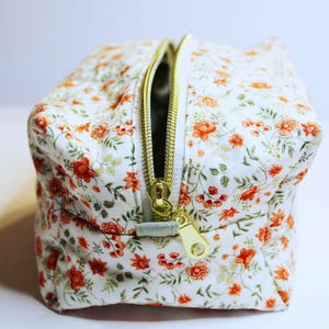 May include: A small, square cosmetic bag with a white background and a floral pattern of orange and red flowers. The bag has a gold zipper and a matching gold zipper pull. The bag is open, revealing the interior.