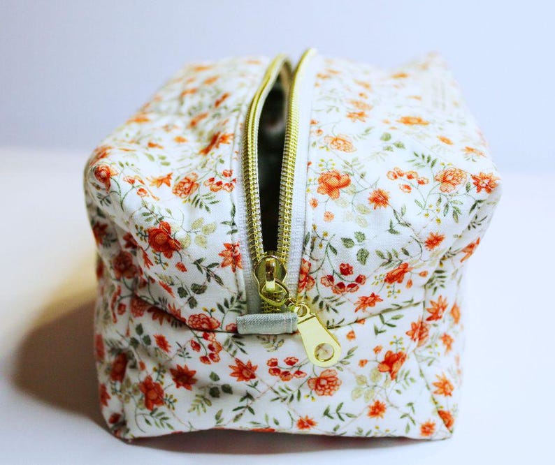 May include: A small, square cosmetic bag with a white background and a floral pattern of orange and red flowers. The bag has a gold zipper and a matching gold zipper pull. The bag is open, revealing the interior.