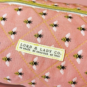 May include: Close-up of a pink quilted fabric with a repeating pattern of yellow and black bees. The fabric has a gold zipper and a rectangular label that reads "LORD & LADY CO. BE BRAVE. BE GRACIOUS. BE TRUE."