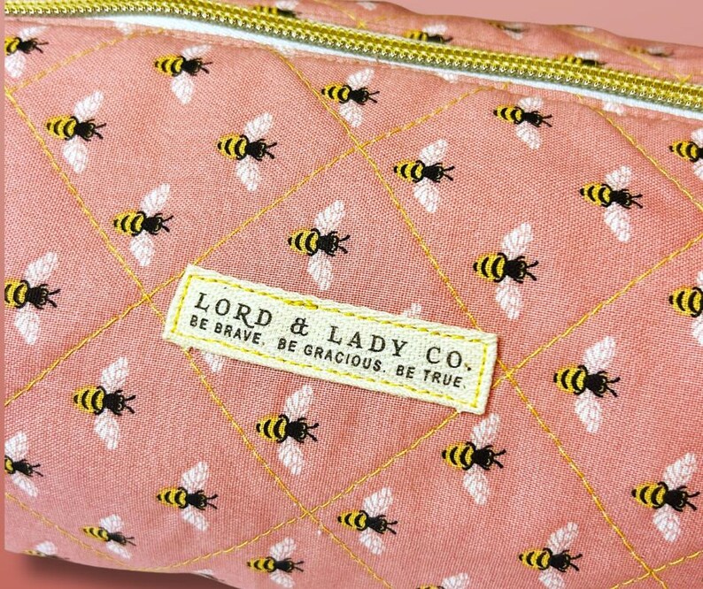 May include: Close-up of a pink quilted fabric with a repeating pattern of yellow and black bees. The fabric has a gold zipper and a rectangular label that reads "LORD & LADY CO. BE BRAVE. BE GRACIOUS. BE TRUE."
