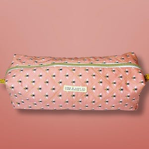 May include: A quilted, rectangular cosmetic bag with a pink background and a pattern of black and yellow bees. The bag has a gold zipper and a small label that reads "LORD & LADY CO."