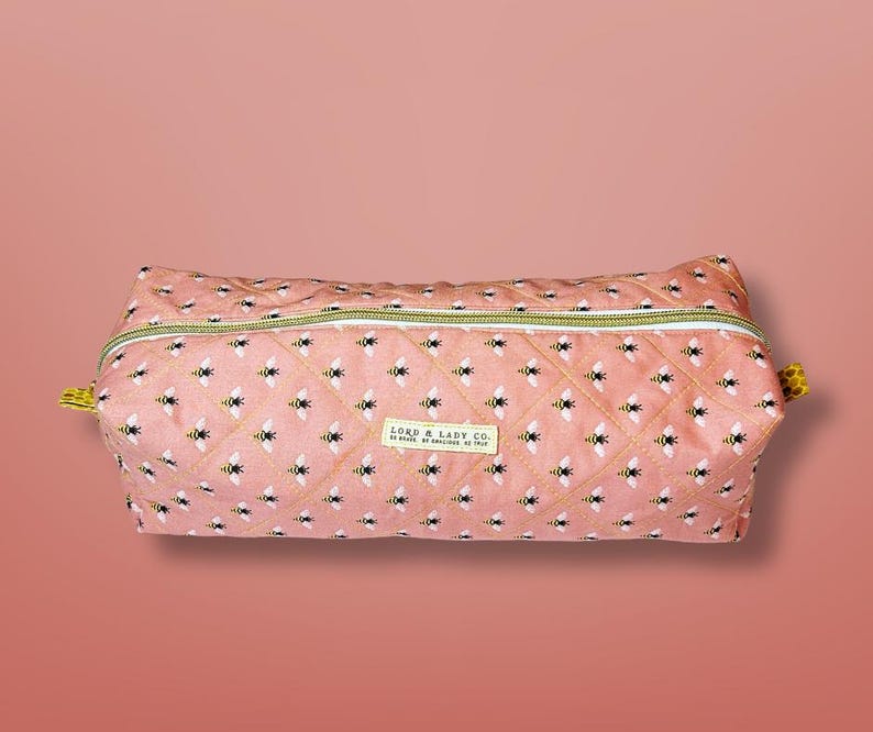 May include: A quilted, rectangular cosmetic bag with a pink background and a pattern of black and yellow bees. The bag has a gold zipper and a small label that reads "LORD & LADY CO."
