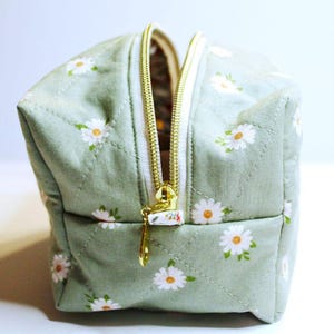 May include: A small, square cosmetic bag with a light green background and a daisy floral pattern. The bag has a gold zipper and a white zipper pull. The bag is quilted with a diamond pattern.