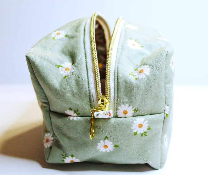 May include: A small, square cosmetic bag with a light green background and a daisy floral pattern. The bag has a gold zipper and a white zipper pull. The bag is quilted with a diamond pattern.