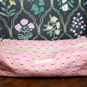 May include: Pink rectangular zippered pouch with a repeating pattern of bees. The pouch has a yellow zipper and a small label. The background is a floral wallpaper with pink, white, and yellow flowers.