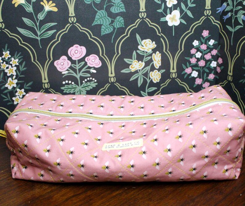 May include: Pink rectangular zippered pouch with a repeating pattern of bees. The pouch has a yellow zipper and a small label. The background is a floral wallpaper with pink, white, and yellow flowers.