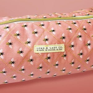 May include: A pink quilted pencil case with a repeating pattern of yellow and black bees. The case has a gold zipper and a rectangular label that reads "LORD & LADY CO. BE BRAVE. BE GRACIOUS. BE TRUE."