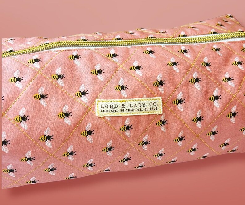 May include: A pink quilted pencil case with a repeating pattern of yellow and black bees. The case has a gold zipper and a rectangular label that reads "LORD & LADY CO. BE BRAVE. BE GRACIOUS. BE TRUE."