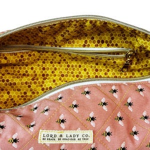 May include: A pink quilted bag with a bee pattern and a gold zipper. The interior features a yellow honeycomb pattern. The bag has a label that reads "LORD & LADY CO. BE BRAVE. BE GRACIOUS. BE TRUE."