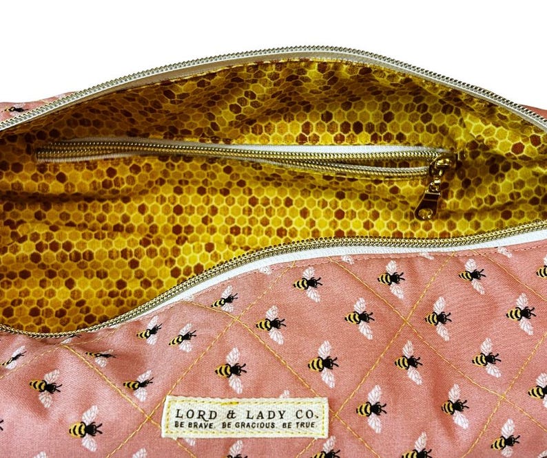 May include: A pink quilted bag with a bee pattern and a gold zipper. The interior features a yellow honeycomb pattern. The bag has a label that reads "LORD & LADY CO. BE BRAVE. BE GRACIOUS. BE TRUE."