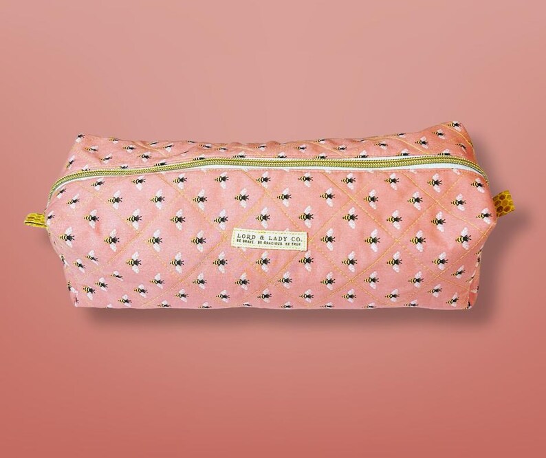 May include: A pink quilted pencil case with a repeating pattern of bees. The case has a yellow zipper and a small label that reads "LORD & LADY CO."