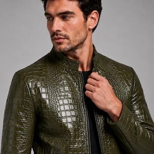 Puede incluir: Una chaqueta de color verde oliva con una textura brillante en relieve que imita la piel de cocodrilo. La prenda cuenta con un cuello alto, una cremallera central negra y bolsillos laterales visibles con cremalleras negras, sobre una camiseta negra.