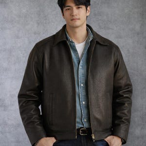 May include: A dark brown leather jacket with a zipper closure and a collared neckline. The jacket has a textured appearance and a classic design. The model is wearing a denim shirt underneath the jacket.